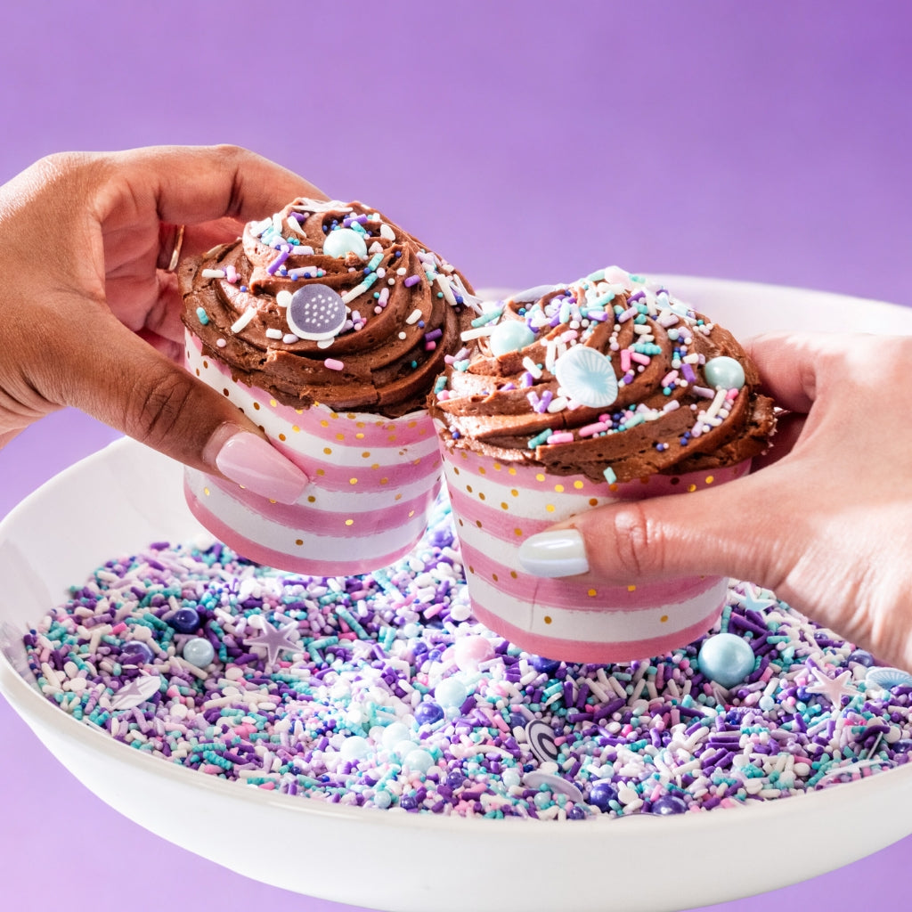 Two chocolate cupcakes with sprinkles held over a bowl of sprinkles