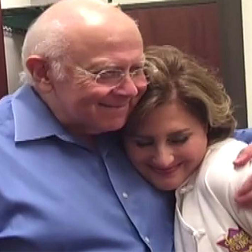 Elderly man in a blue shirt hugging a woman in a white coat.